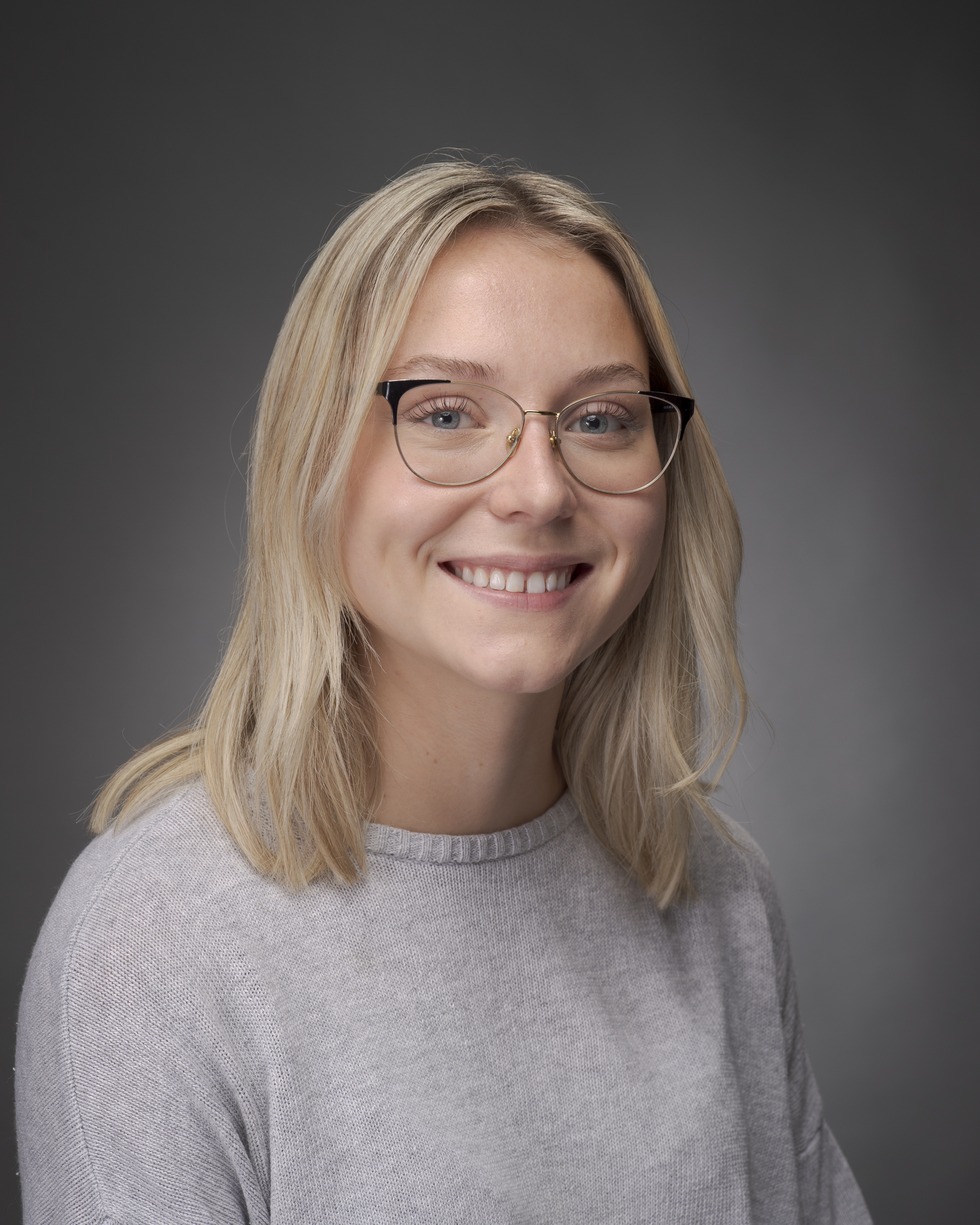 Photo of white woman with blond hair and a white shirt on. The woman is smiling. The background is gray.