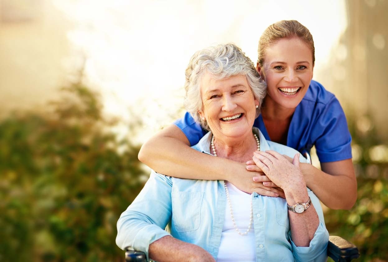 Caregiving Hug A smiling older female in a light blue button-down shirt over a white shirt sitting with a smiling younger female wearing a medium blue shortsleeved shirt bent hugging the older female around the neck from behind.