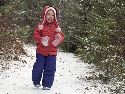 Physical activity doesn't have to be structure to be beneficial. A small child in a colorful snowsuit runs down a snowcovered trail in the pine trees.