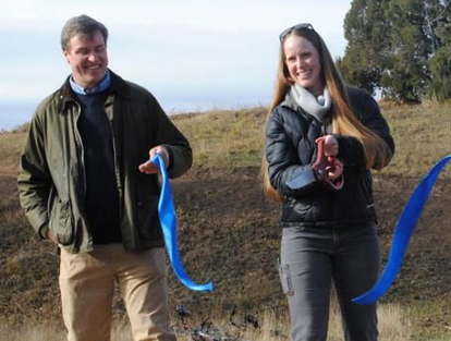 A woman smiles while she cuts a large blue ribbon with an oversized pair of scissors